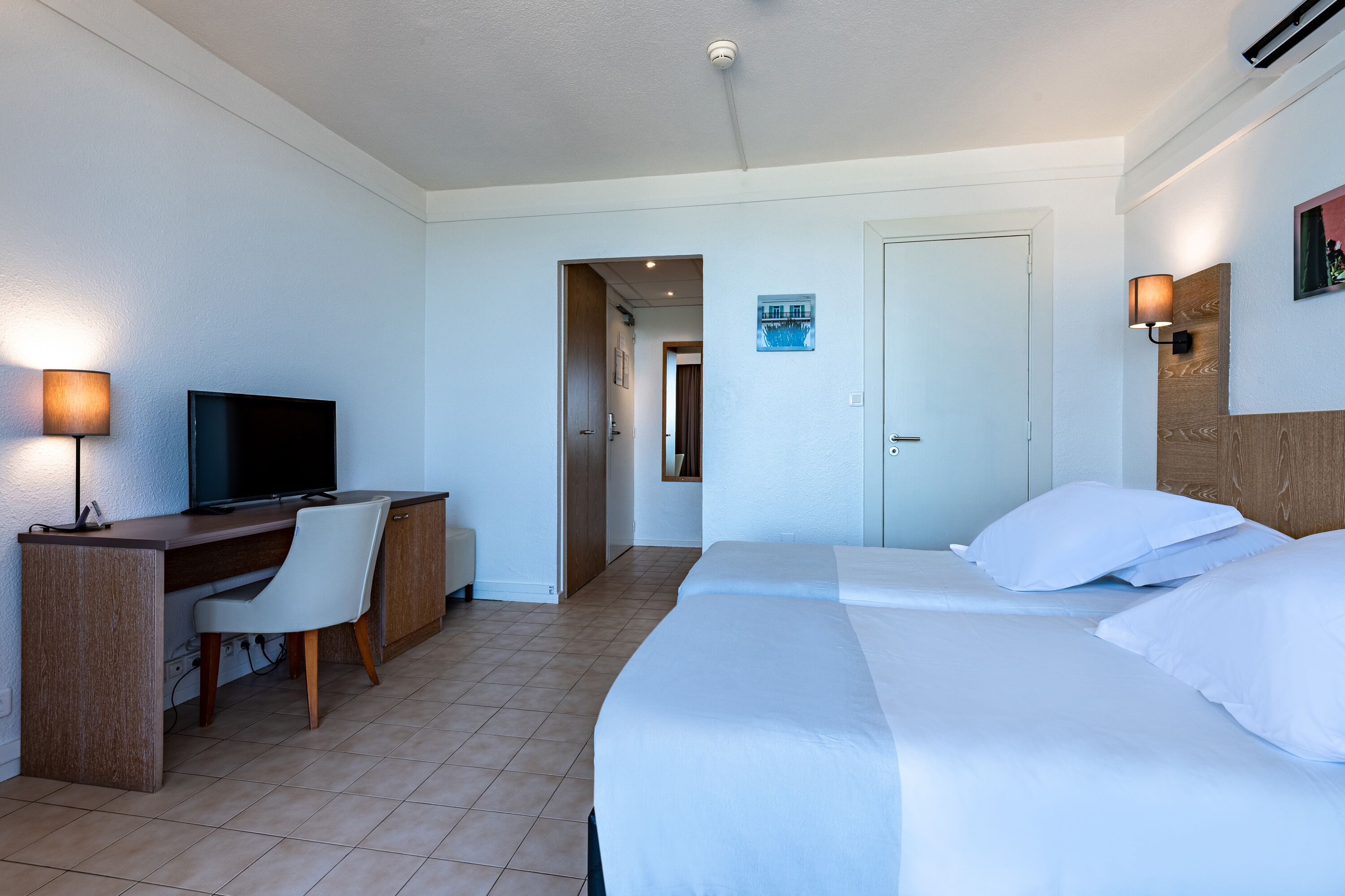 A hotel room with a bed, a desk with a chair, a television, and a wall-mounted lamp.