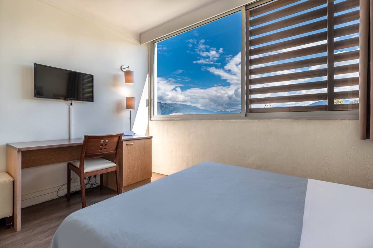 Une chambre d’hôtel équipée d’un lit, d’un bureau, d’une chaise, d’une télévision et d’une grande fenêtre avec des stores.
