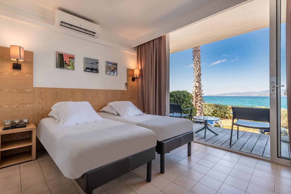 Une chambre d’hôtel avec deux lits, une tête de lit en bois, des œuvres d’art accrochées au mur et un balcon avec vue sur la mer.
