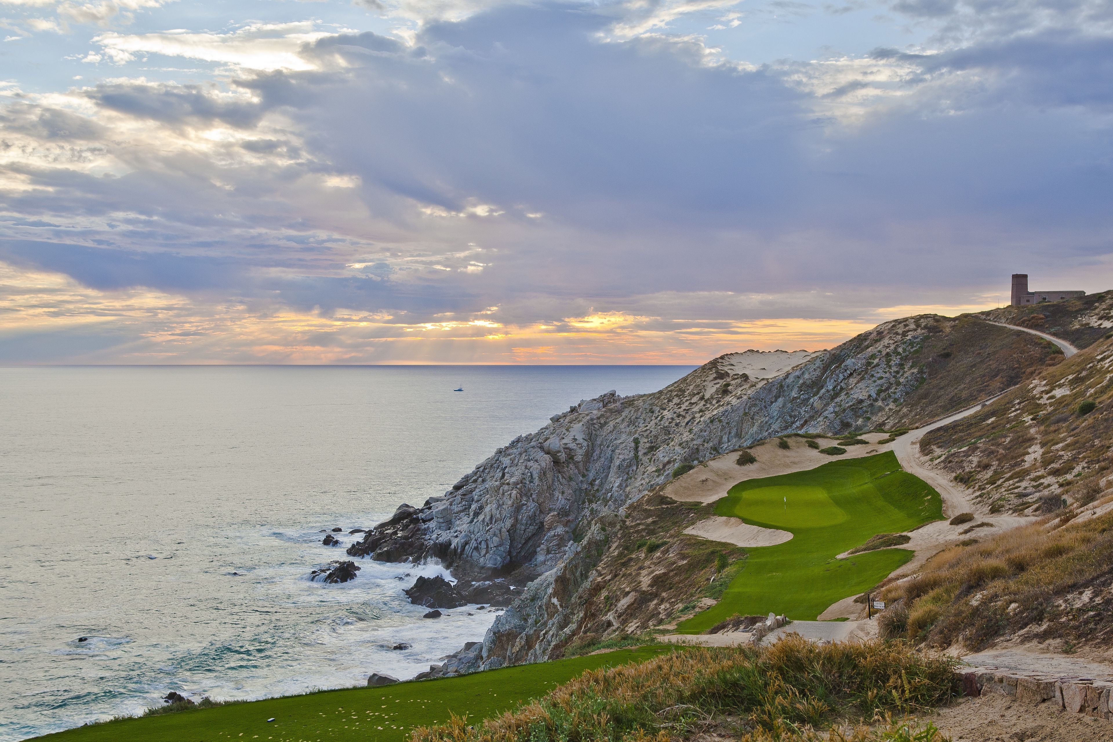 A coastal golf course with a clear path, rocky cliffs, and a distant building.