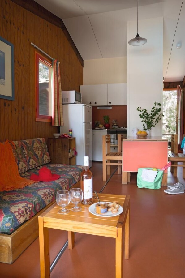 A cozy living room with a sofa, a wooden table with a plate of food and glasses, a kitchen area with a refrigerator and a microwave, and a dining area with a table and chairs. - Sowell LES Sentes (Carcans)