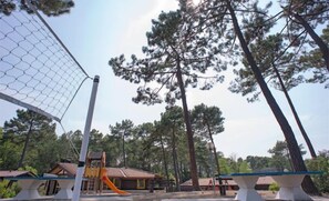 A playground with a slide and a basketball hoop, surrounded by tall trees and a building with a tiled roof. - Sowell LES Sentes (Carcans)