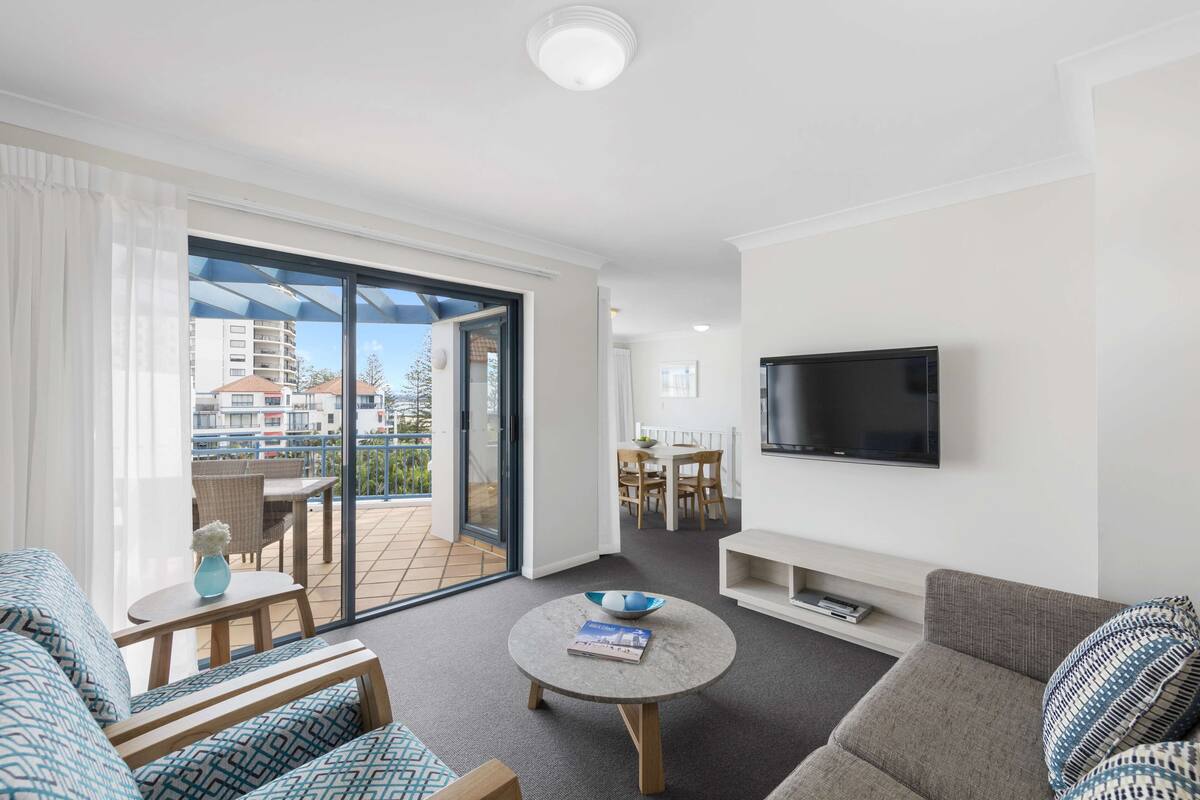 A modern living room with a sofa, a coffee table, and a TV. There is a balcony with a view of buildings.