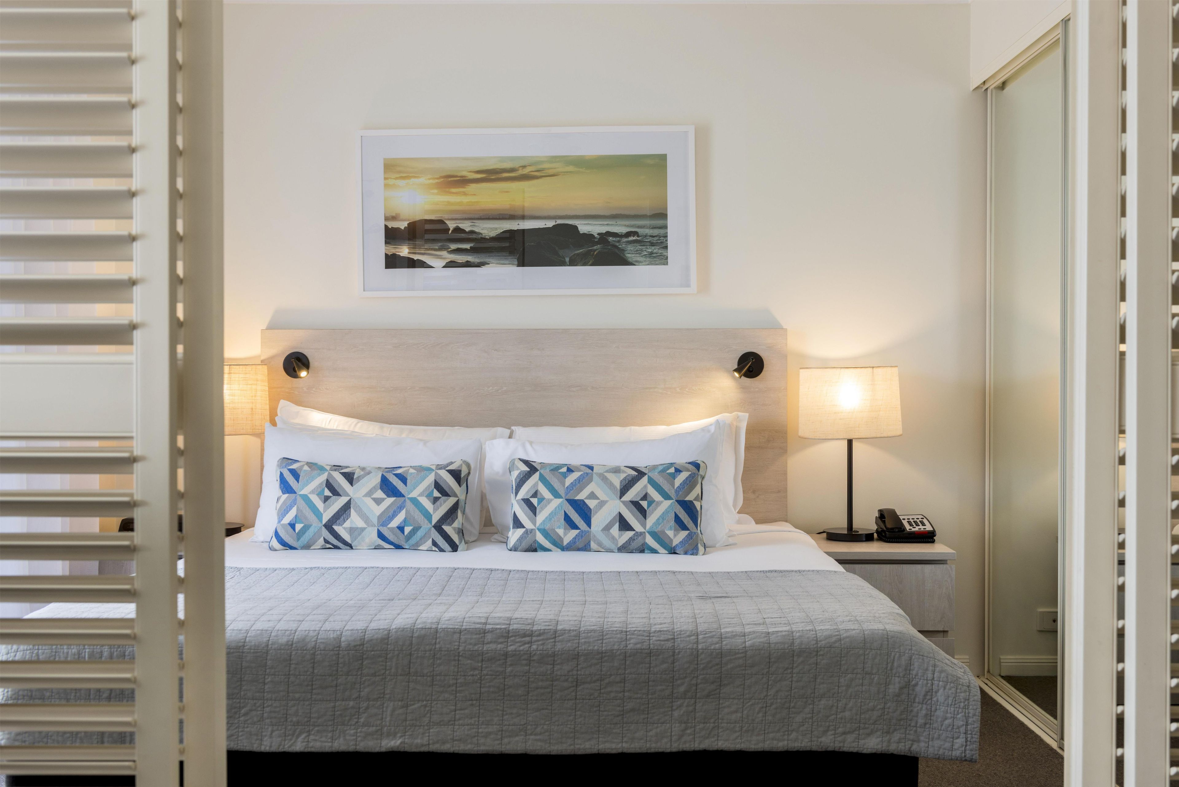 A neatly made bed with a geometric patterned pillow, a bedside lamp, and a framed picture of a sunset over the ocean on the wall.