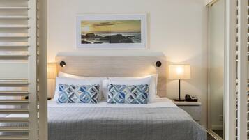 A neatly made bed with a geometric patterned pillow, a bedside lamp, and a framed picture of a sunset over the ocean on the wall.