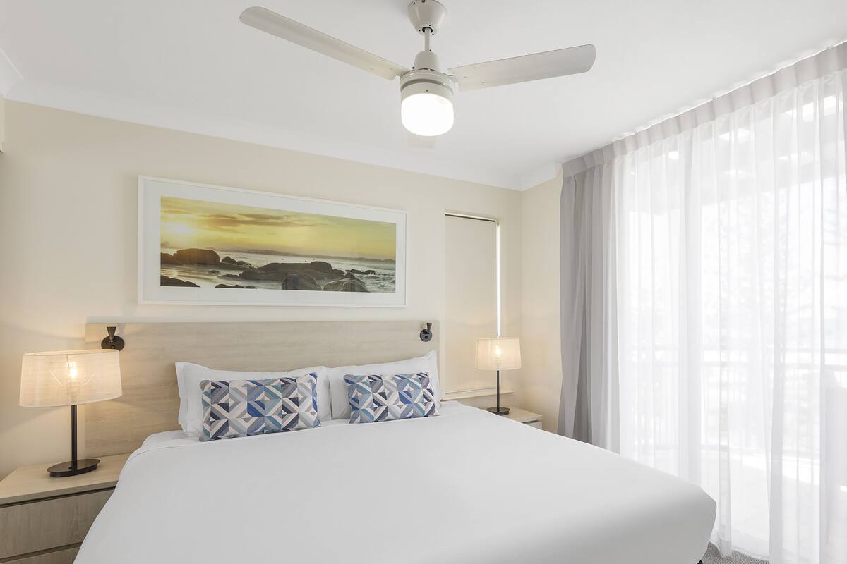 A hotel room with a large bed, two bedside lamps, a ceiling fan, a painting on the wall, and sheer curtains on the window.