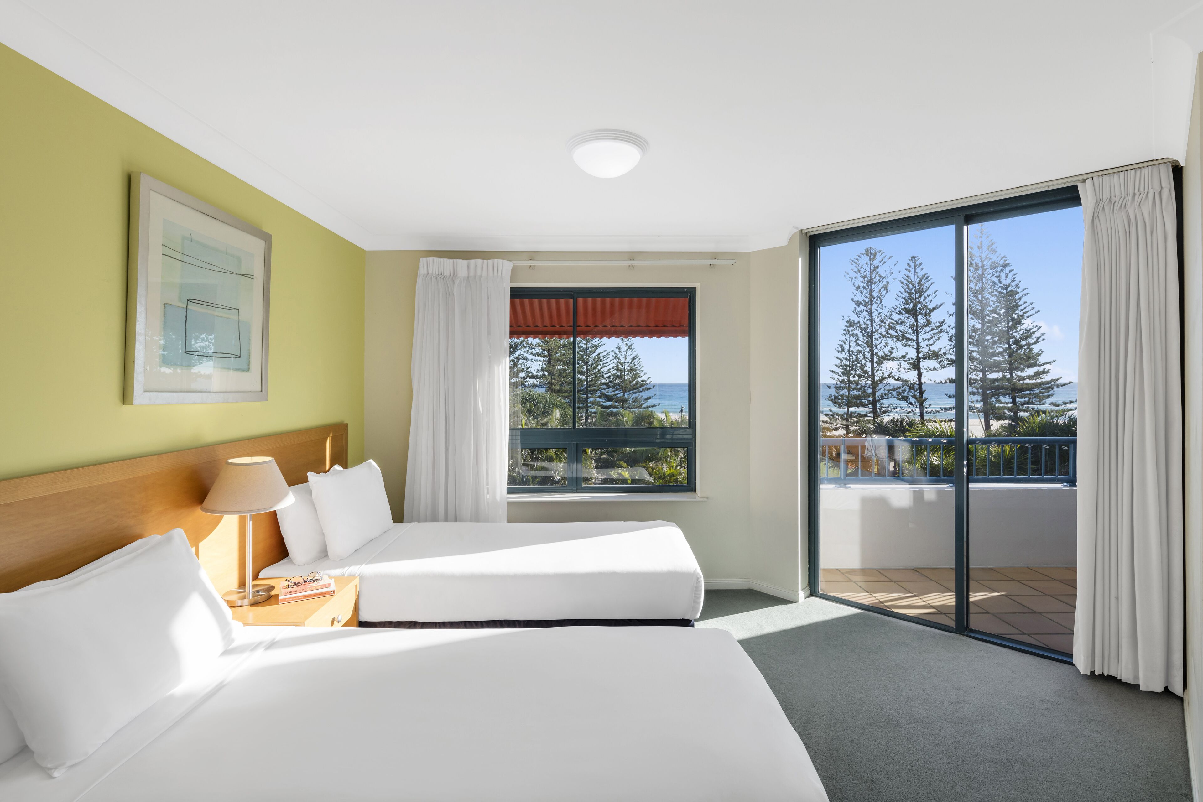 A hotel room with two beds, a large window with a view of trees and the sea, and a balcony with curtains.