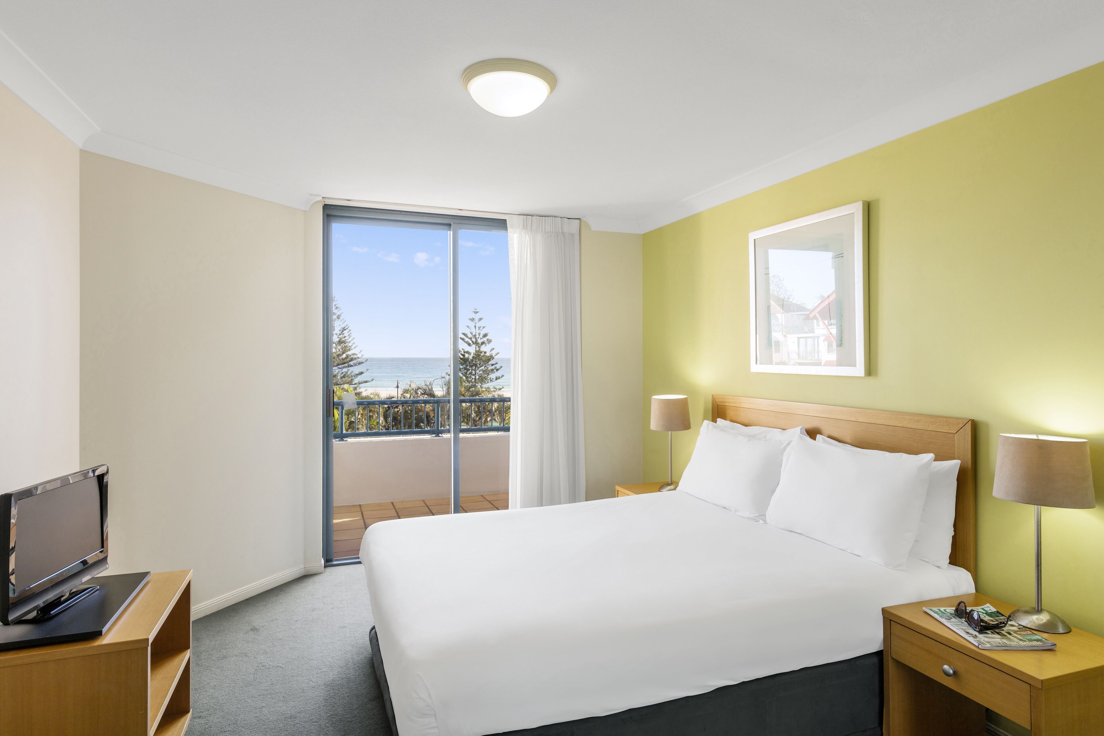 A hotel room with a large bed, a TV on a stand, a nightstand with a lamp, and a view of the sea through a balcony.