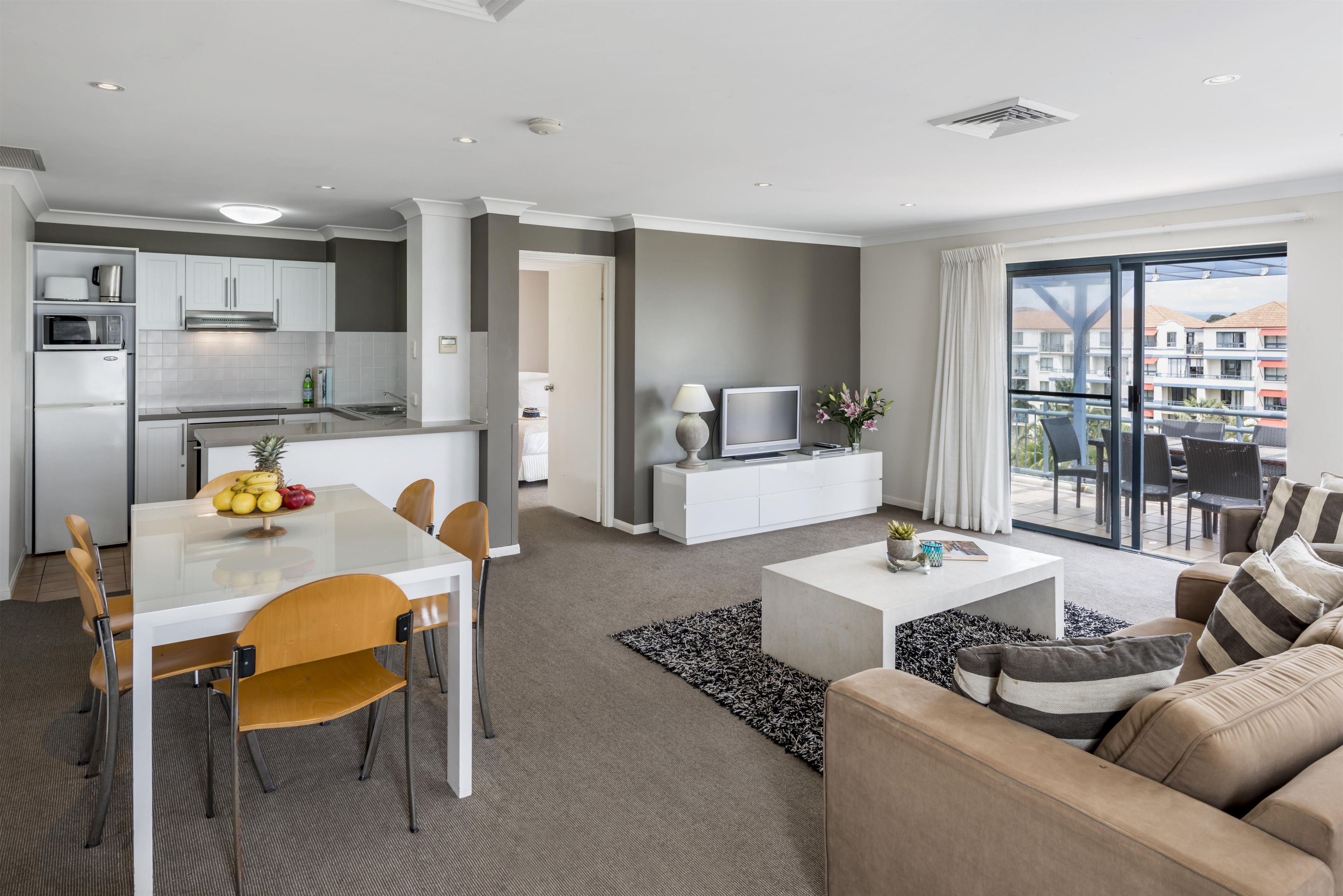 A modern hotel room with a dining area, kitchenette, living room, and a balcony with a city view.