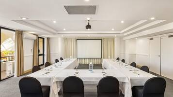 A conference room with a long table, chairs, a projector screen, and a large window with curtains.
