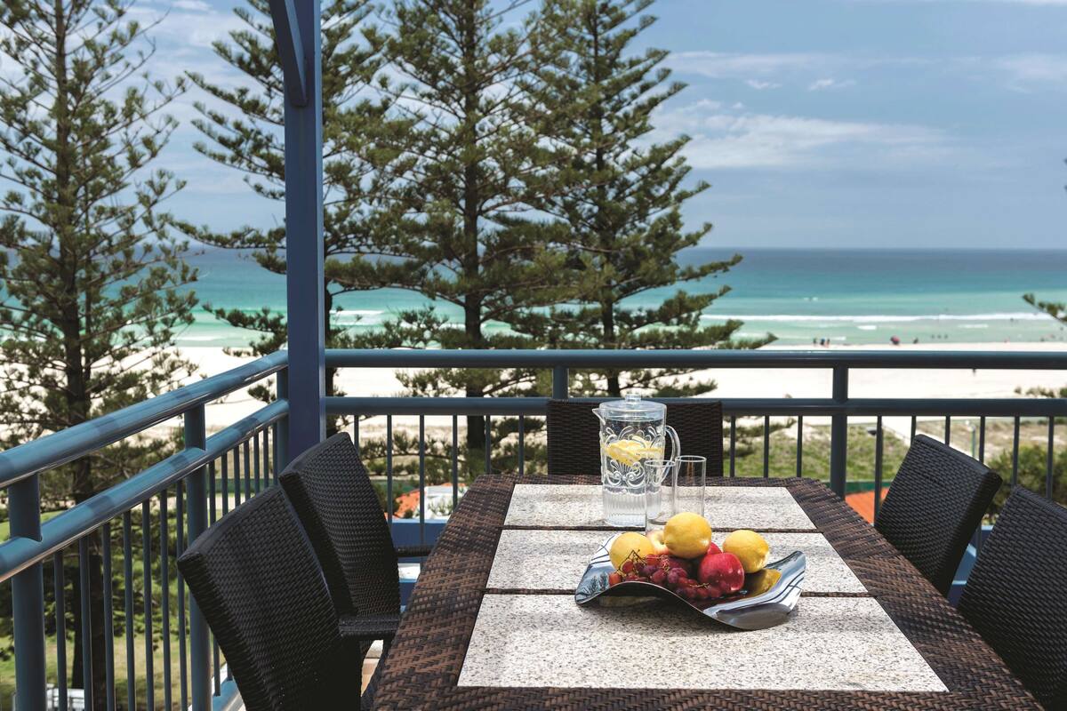 A table with a fruit bowl, pitcher, and glasses on a balcony overlooking the beach.