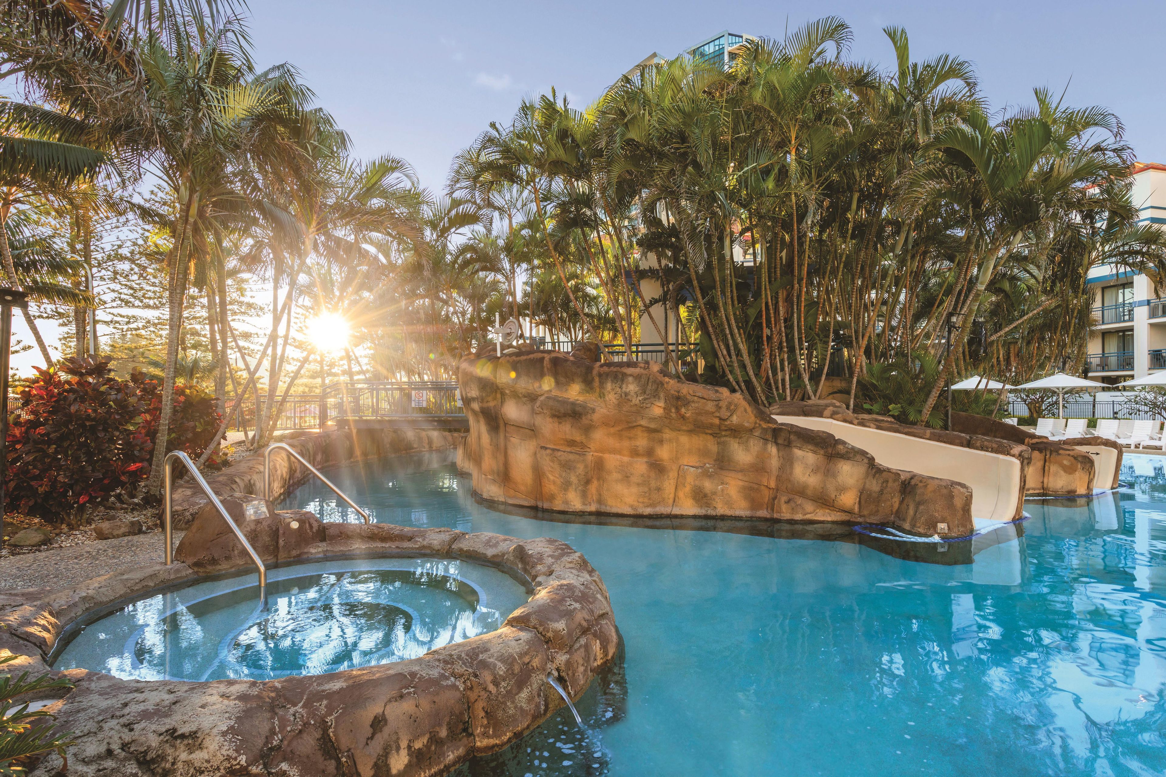 A pool area with a hot tub, surrounded by palm trees and a building in the background.