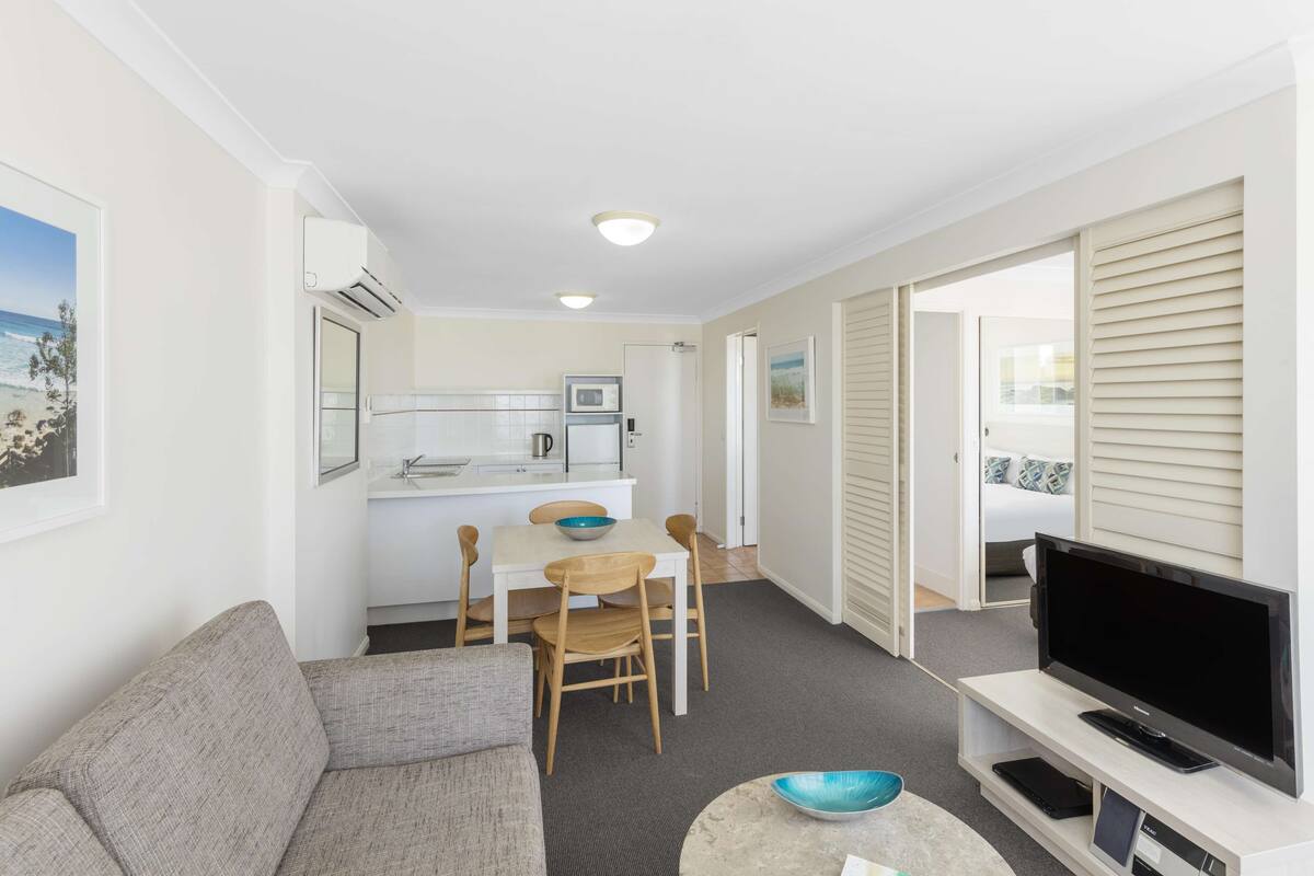 A compact living space with a kitchenette, a sofa, a dining table with chairs, a flat-screen TV, and a large window with a view of the beach.