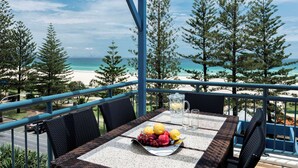 A table with a fruit bowl, pitcher, and glasses, overlooking a beach and trees.
