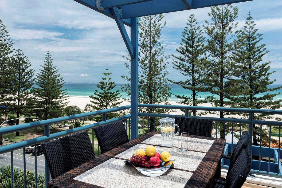 A table with a fruit bowl, pitcher, and glasses, overlooking a beach and trees.