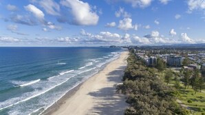 Vista costera con playa arenosa, olas del océano y un paisaje urbano con edificios y vegetación.