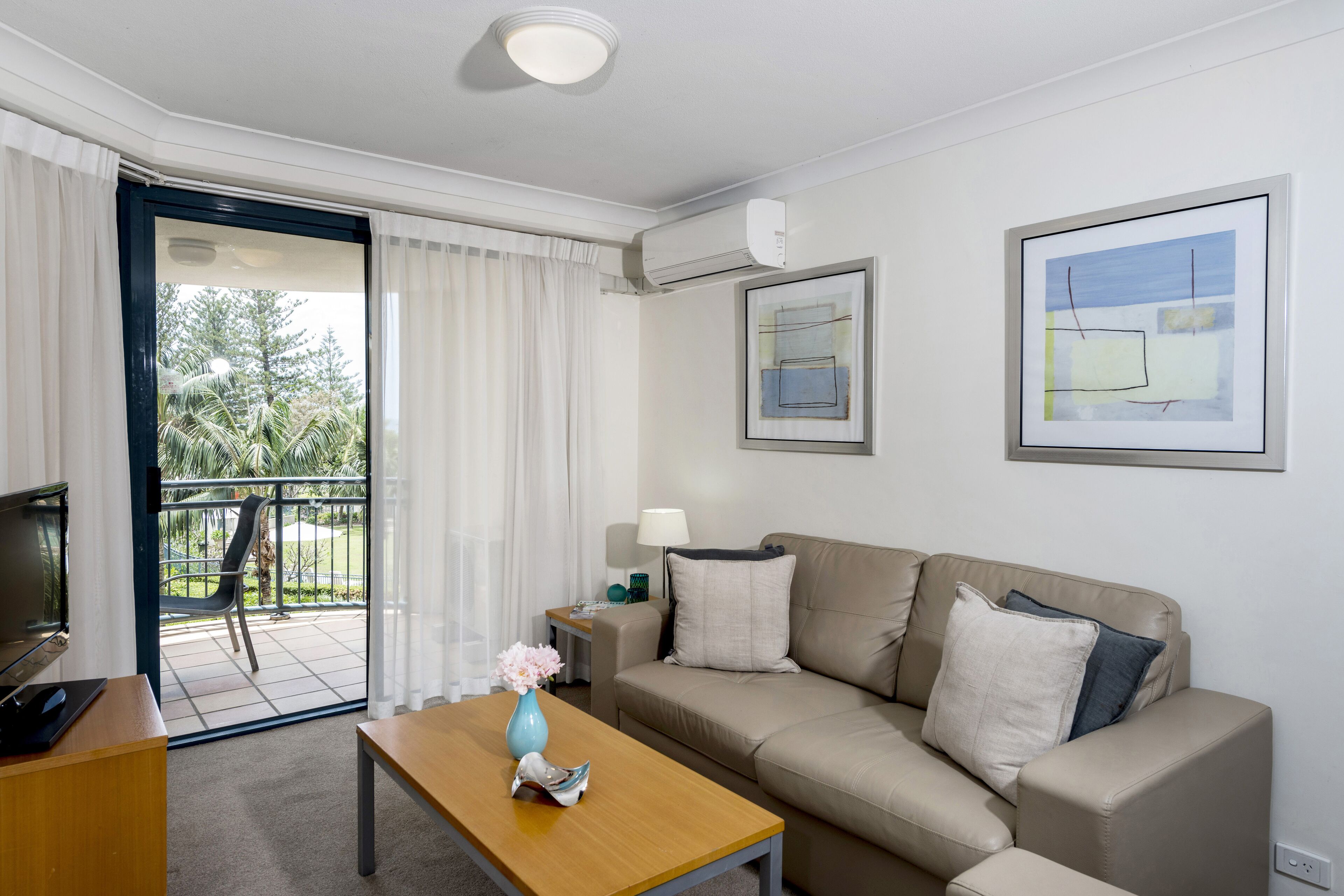 A modern hotel room with a beige sofa, a wooden coffee table with a vase, a TV, and a balcony with a view of trees.