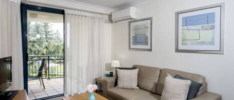 Quarto de hotel moderno com um sofá bege, uma mesa de centro de madeira com um vaso, uma TV e uma varanda com vista para árvores.