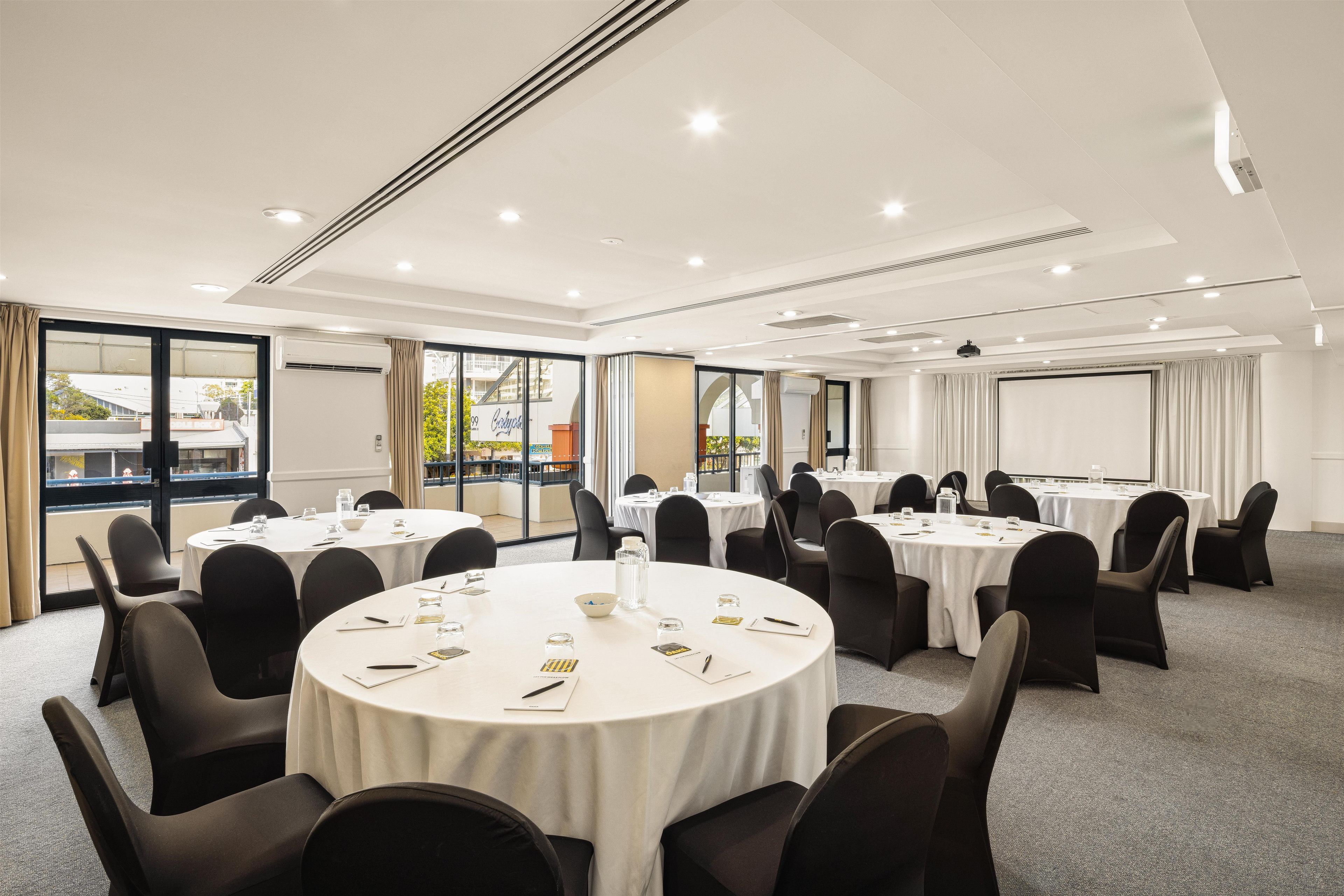 A conference room with multiple round tables, black chairs, and a large projection screen.