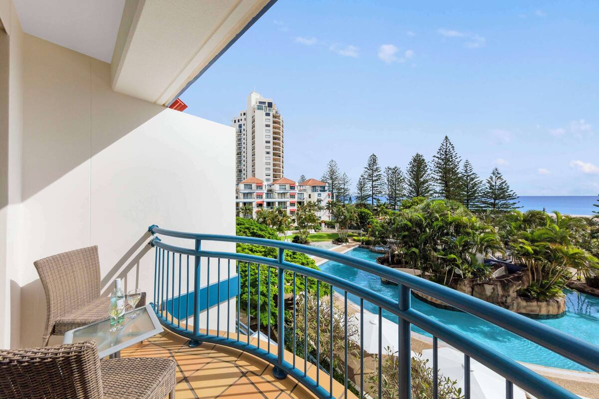 A balcony with wicker furniture, a glass table, and a pool area with a blue railing.