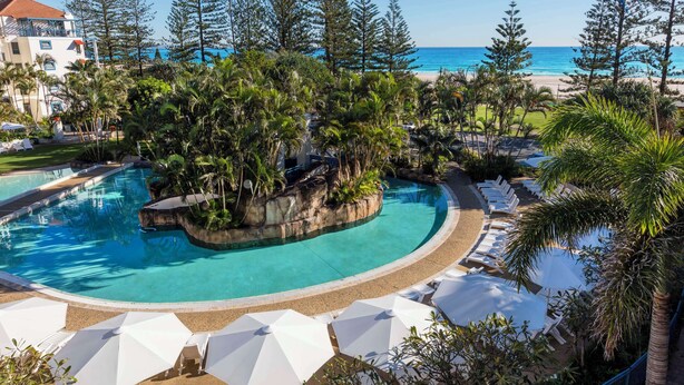 A pool with a natural rock island, surrounded by lounge chairs and umbrellas, with a beach and ocean view in the background.