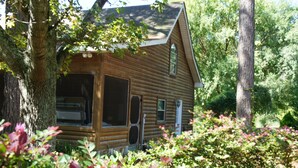 A rustic log cabin with a large front window and a door, surrounded by dense foliage. - Shalom House Bed and Breakfast (Tifton)
