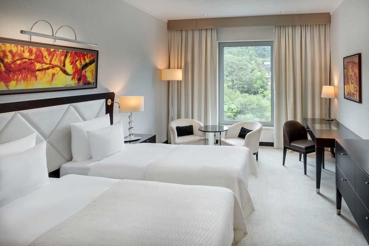 A hotel room with two beds, a desk, a chair, and a view of trees through the window.