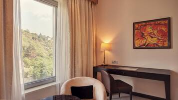 A hotel room with a desk, chair, and a view of a forest through a window.