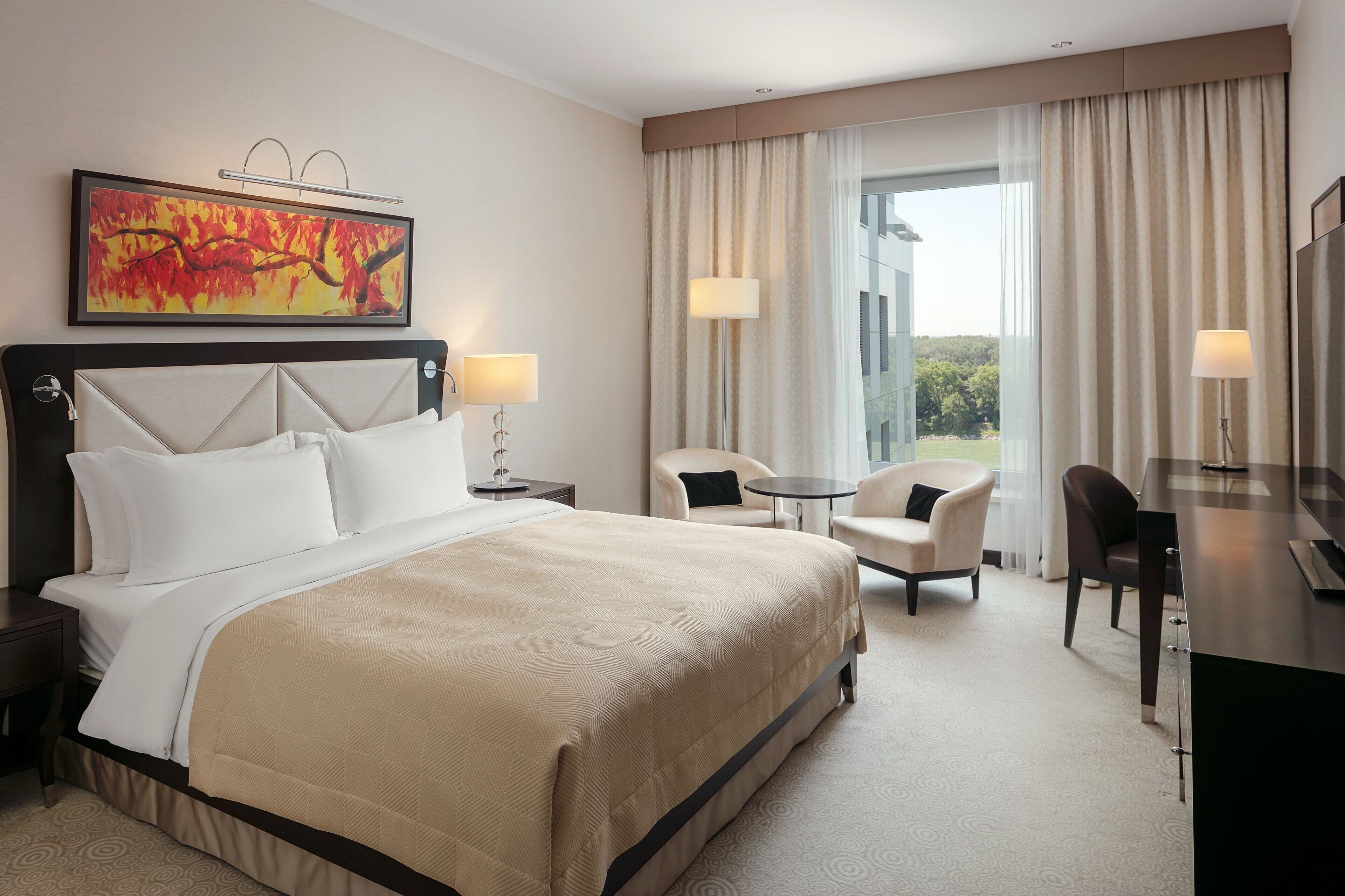 A hotel room with a large bed, two bedside tables, a desk, a chair, and a large window with curtains.