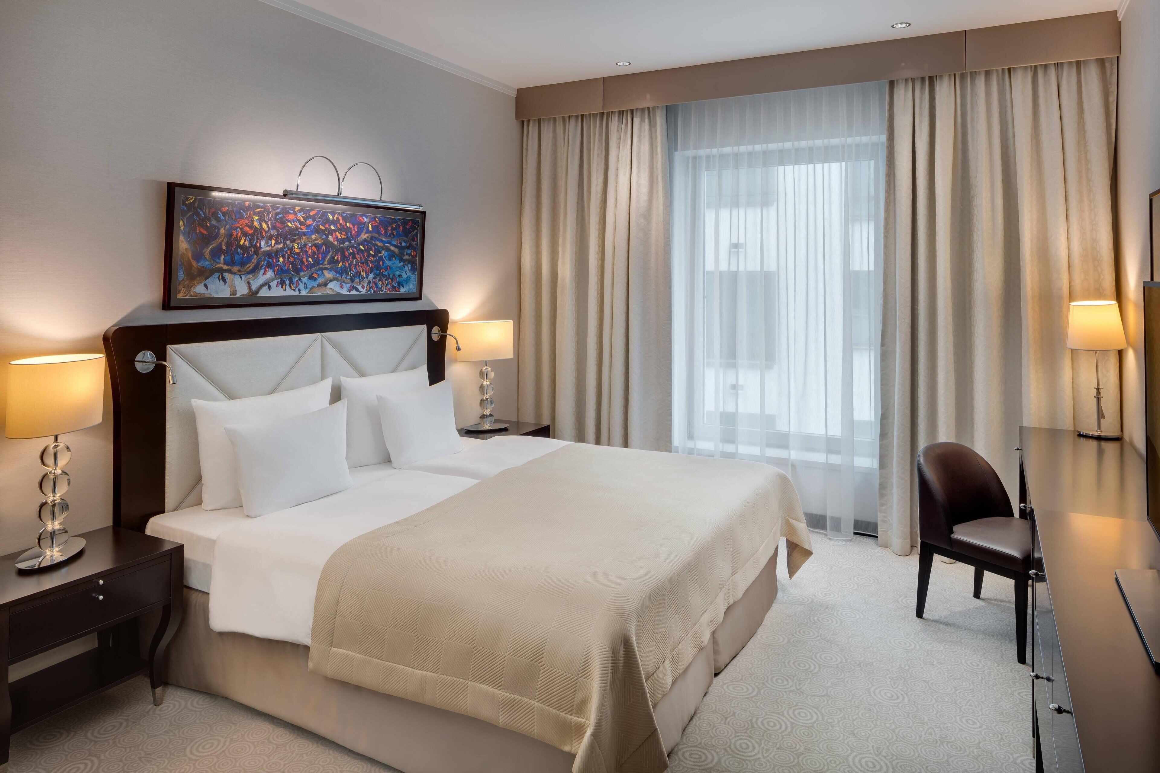 A hotel room with a large bed, two bedside lamps, a desk with a chair, a painting above the bed, and a window with curtains.