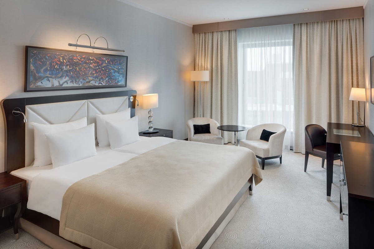 A hotel room with a large bed, two bedside tables, a desk, a chair, and a small table. There is a painting on the wall and a window with curtains.