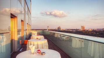 A rooftop terrace with tables set for dining, overlooking a cityscape with buildings and a tower.