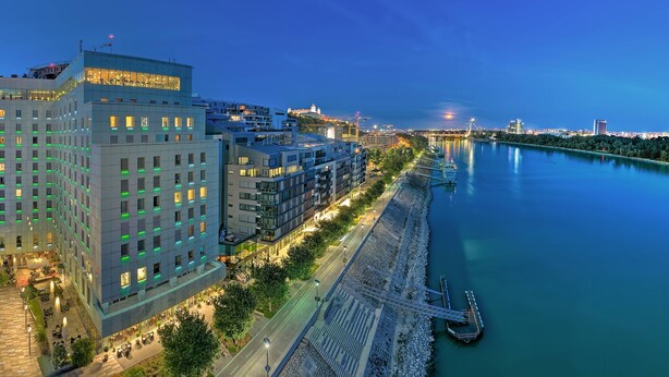 Een modern hotel met uitzicht op een rivier, een aanlegsteiger en een brug op de achtergrond.