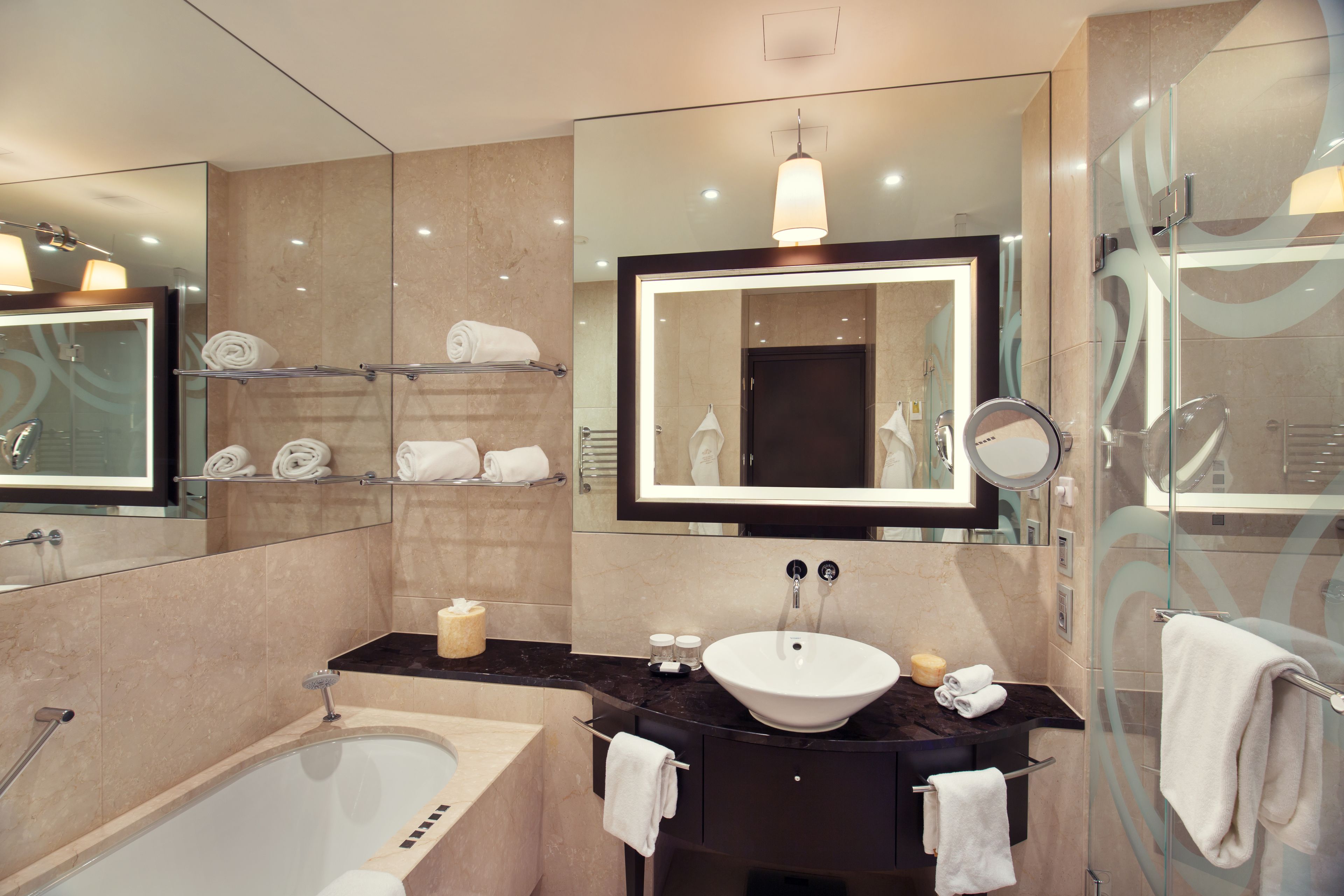 A modern bathroom with a bathtub, a sink, and a large mirror. There are towels on a shelf and a glass shower enclosure.
