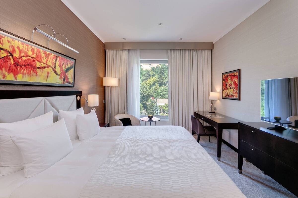 A modern hotel room with a large bed, a desk, and a view of greenery outside.
