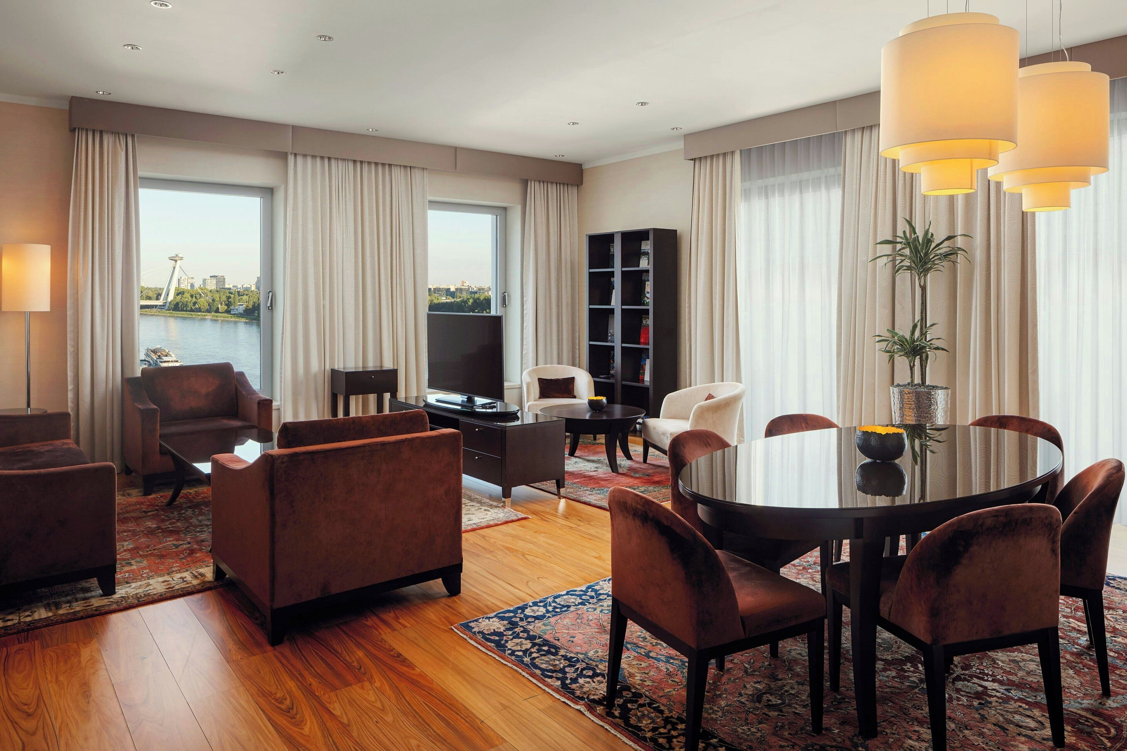 A modern hotel room with a large window, a round dining table, and comfortable seating.