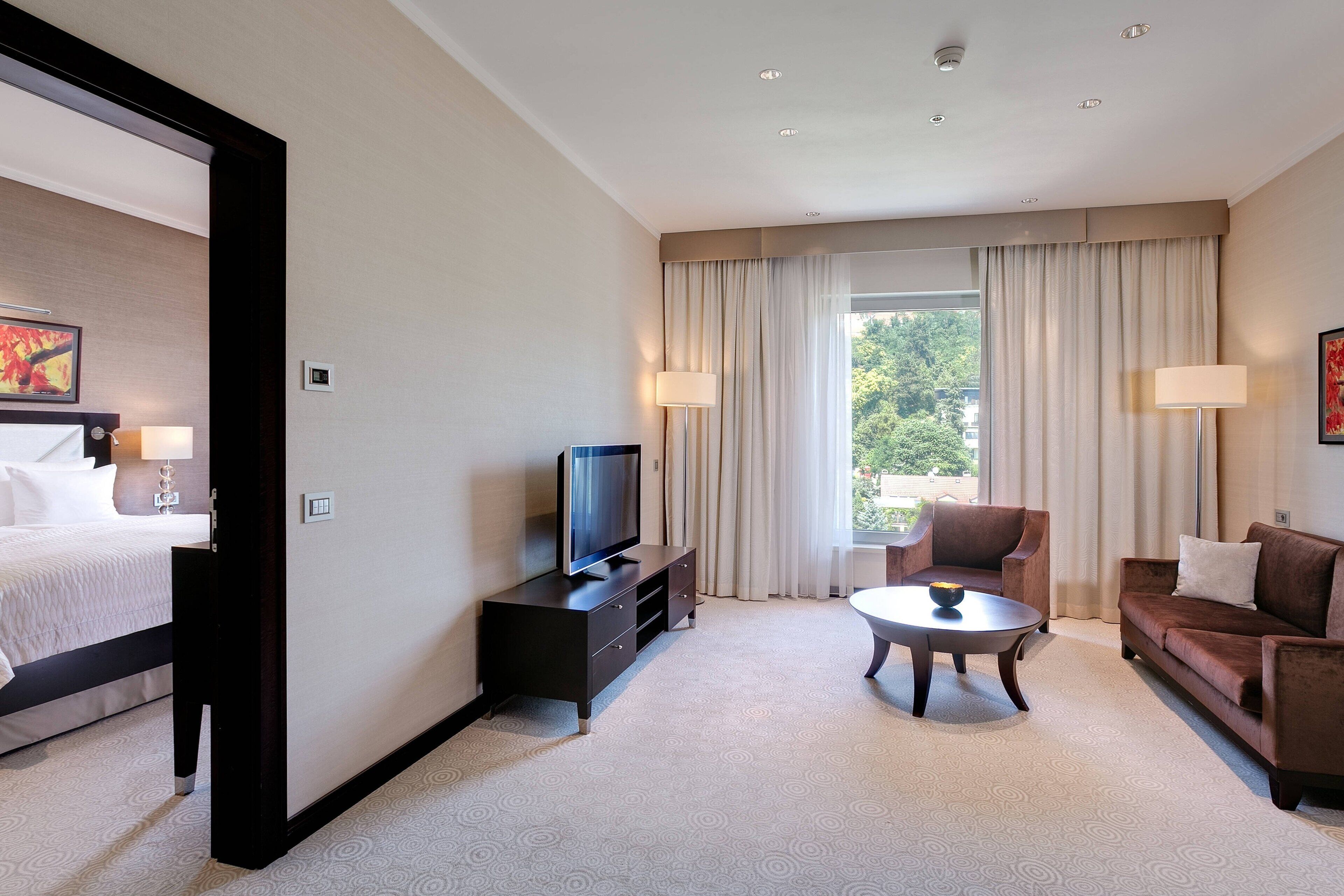 A hotel room with a bed, a TV on a stand, a sofa, a coffee table, and a window with curtains.