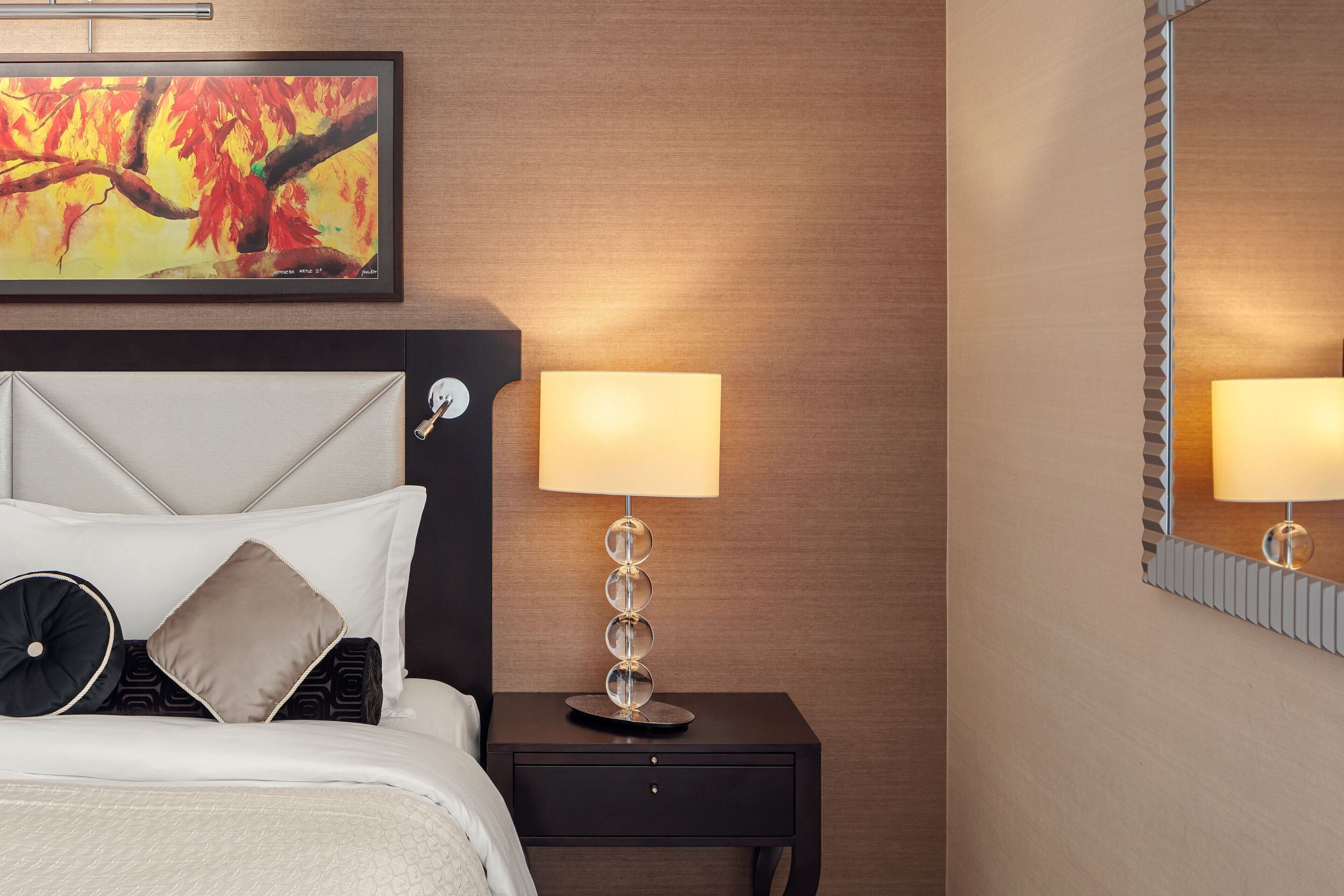 A neatly made bed with a headboard, a bedside table with a lamp, and a wall-mounted painting.