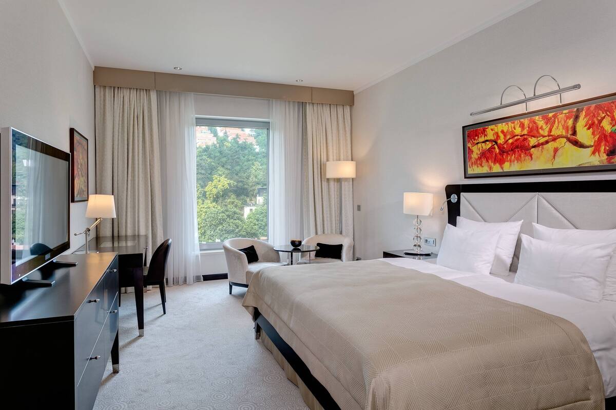 A hotel room with a large bed, a desk with a TV, a chair, a small table, and a window with curtains.