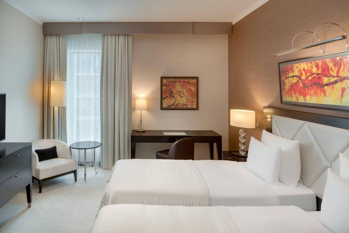 A hotel room with two beds, a desk, a chair, a TV, and a painting on the wall.