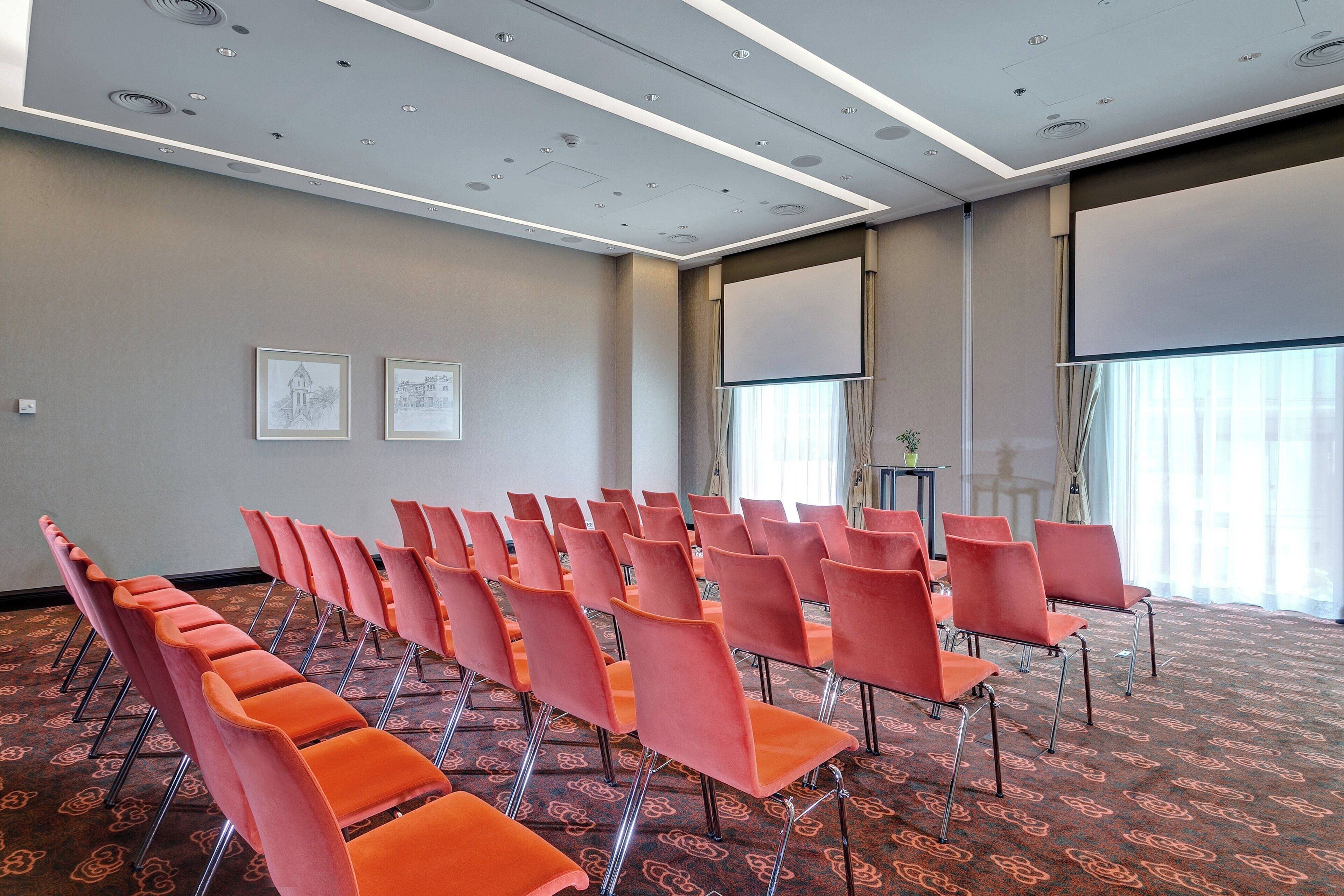 A conference room with rows of orange chairs, a large screen, and a patterned carpet.