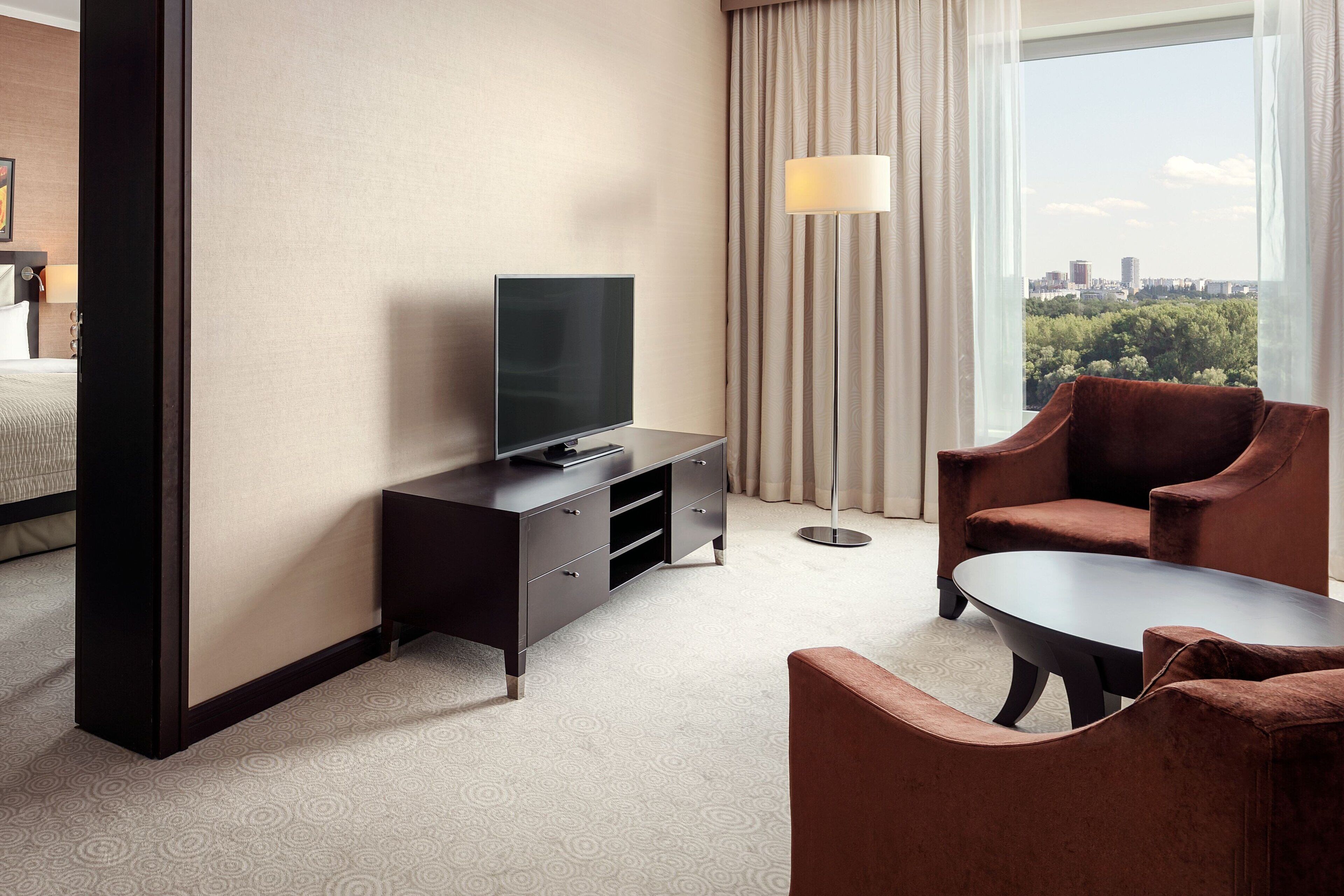 A hotel room with a bed, a TV on a dresser, a sofa, a coffee table, and a view of a cityscape through large windows.