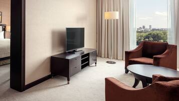 A hotel room with a bed, a TV on a dresser, a sofa, a coffee table, and a view of a cityscape through large windows.