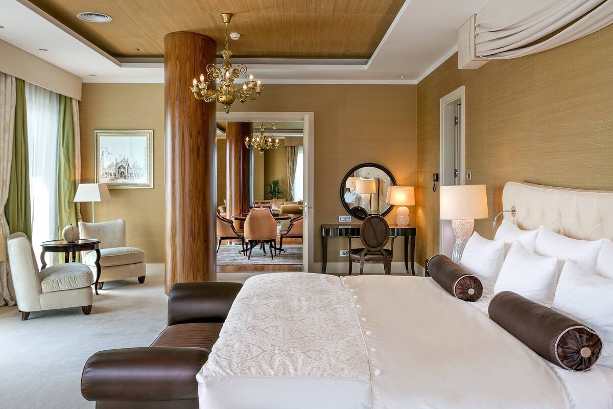 A hotel room with a large bed, two armchairs, a dining area in the background, and a chandelier.