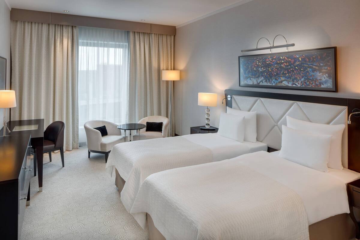 A hotel room with two beds, a desk, a chair, a small table, and a painting on the wall.