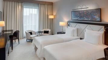 A hotel room with two beds, a desk, a chair, a small table, and a painting on the wall.