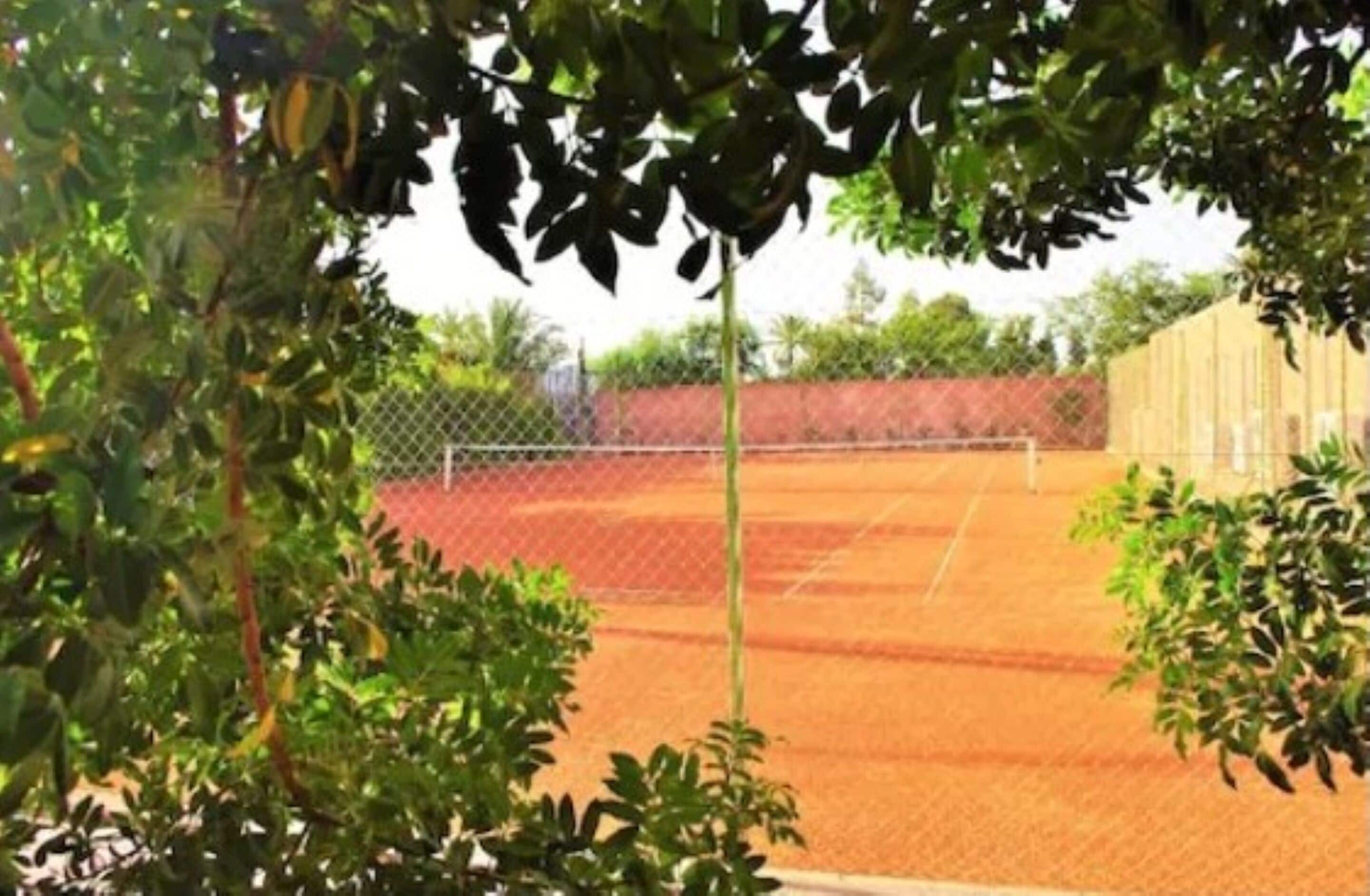 A tennis court with a chain-link fence, surrounded by greenery and a building in the background.
