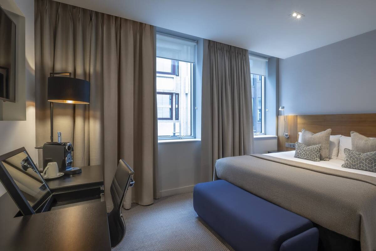 A hotel room with a large bed, a desk with a lamp, a chair, and a window with curtains.