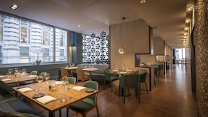 Een restaurant met houten tafels en groene stoelen, grote ramen en een muren met patroon.