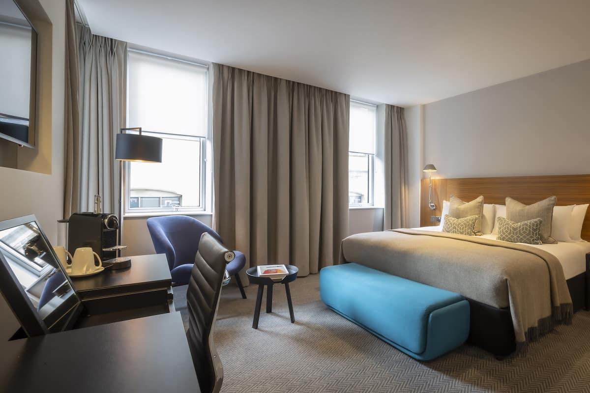 A hotel room with a bed, a desk with a coffee maker, a chair, a small table, and a blue ottoman.